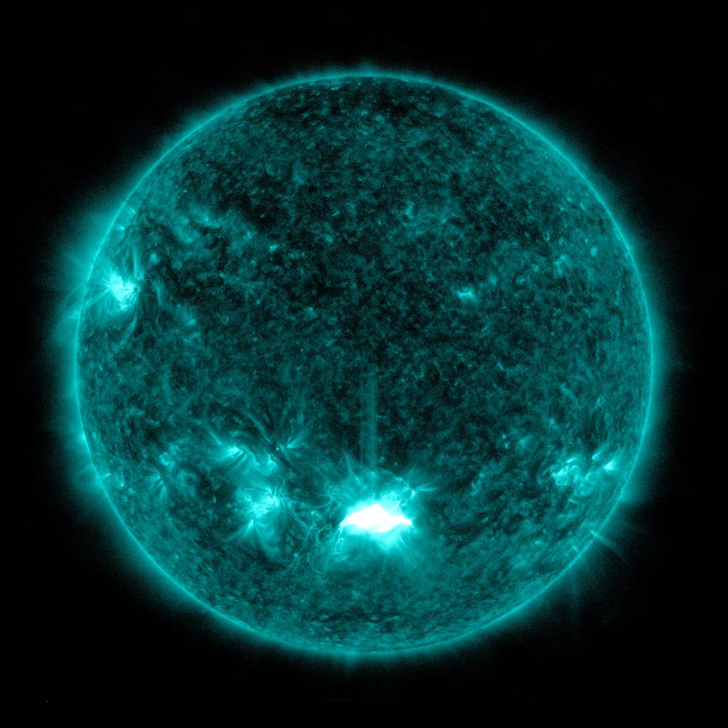 A satellite image of the Sun, colored in teal, shows a bright flare at the Sun's lower center.